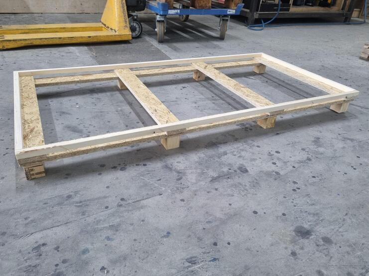 Holzpalette auf einem betonierten Werkstattboden, mit sichtbaren Holzverbindungen und -oberfl&auml;che.