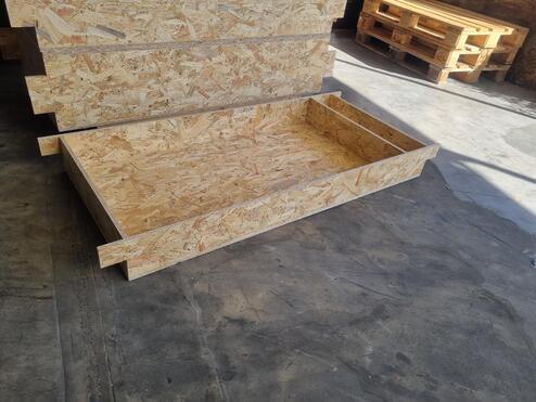 Holzrahmen aus OSB-Platten liegt auf einem Betonboden, umgeben von gestapelten Holzpaletten.