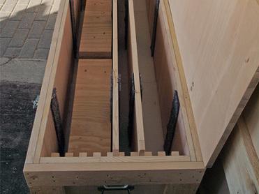 Offene Holzbox mit mehreren Fächern, gefüllt mit Holzplatten, auf einem gepflasterten Boden.